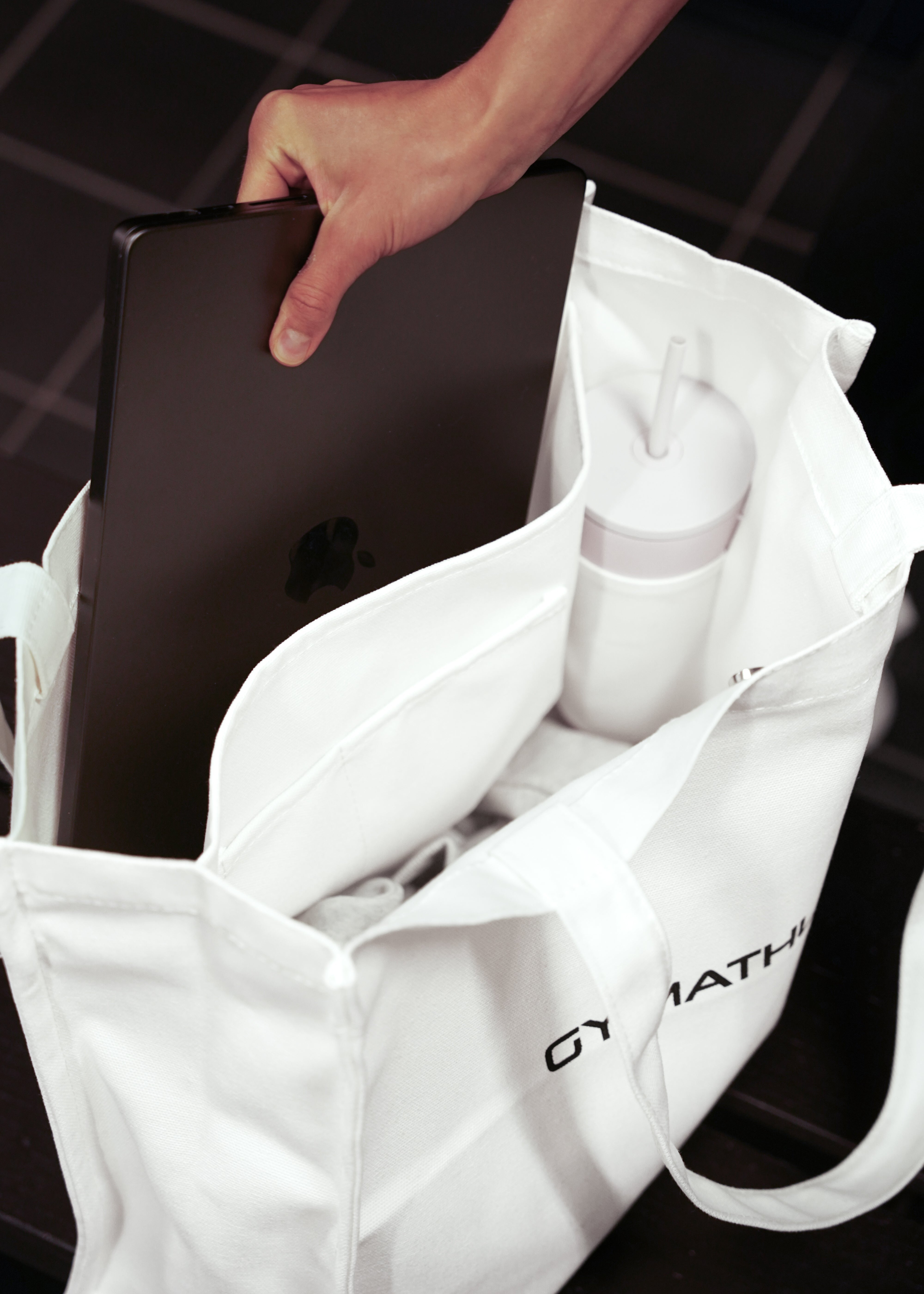 Person holding a black laptop inside a white tote bag with 'GYMATHLETE' branding.
White tote bag with 'GYMATHLETE' branding on a light gray background. Balance Tote.