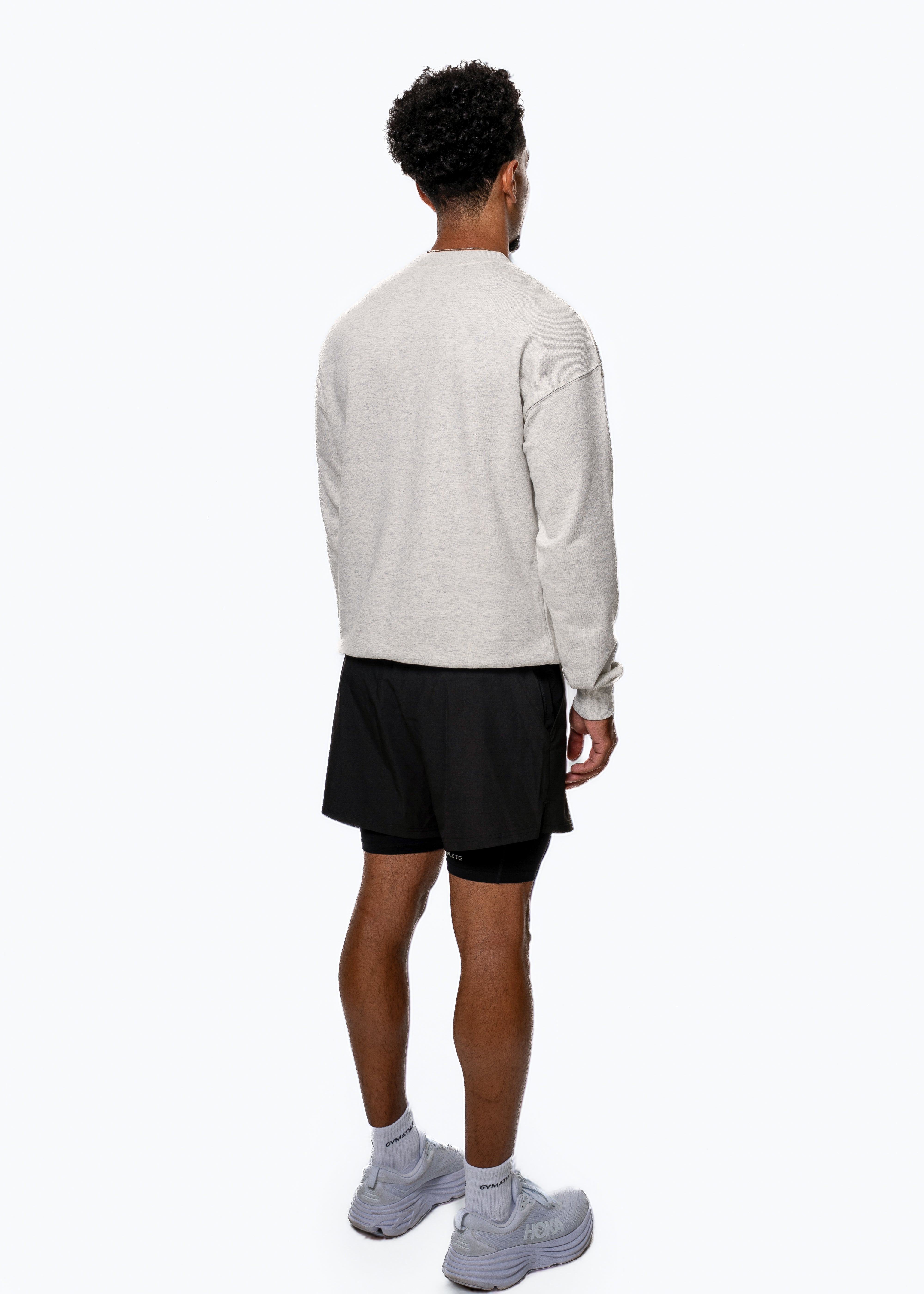 Person wearing a light gray sweater, black shorts, and white sneakers on a white background. Elevate Sweatshirt, Gymathlete