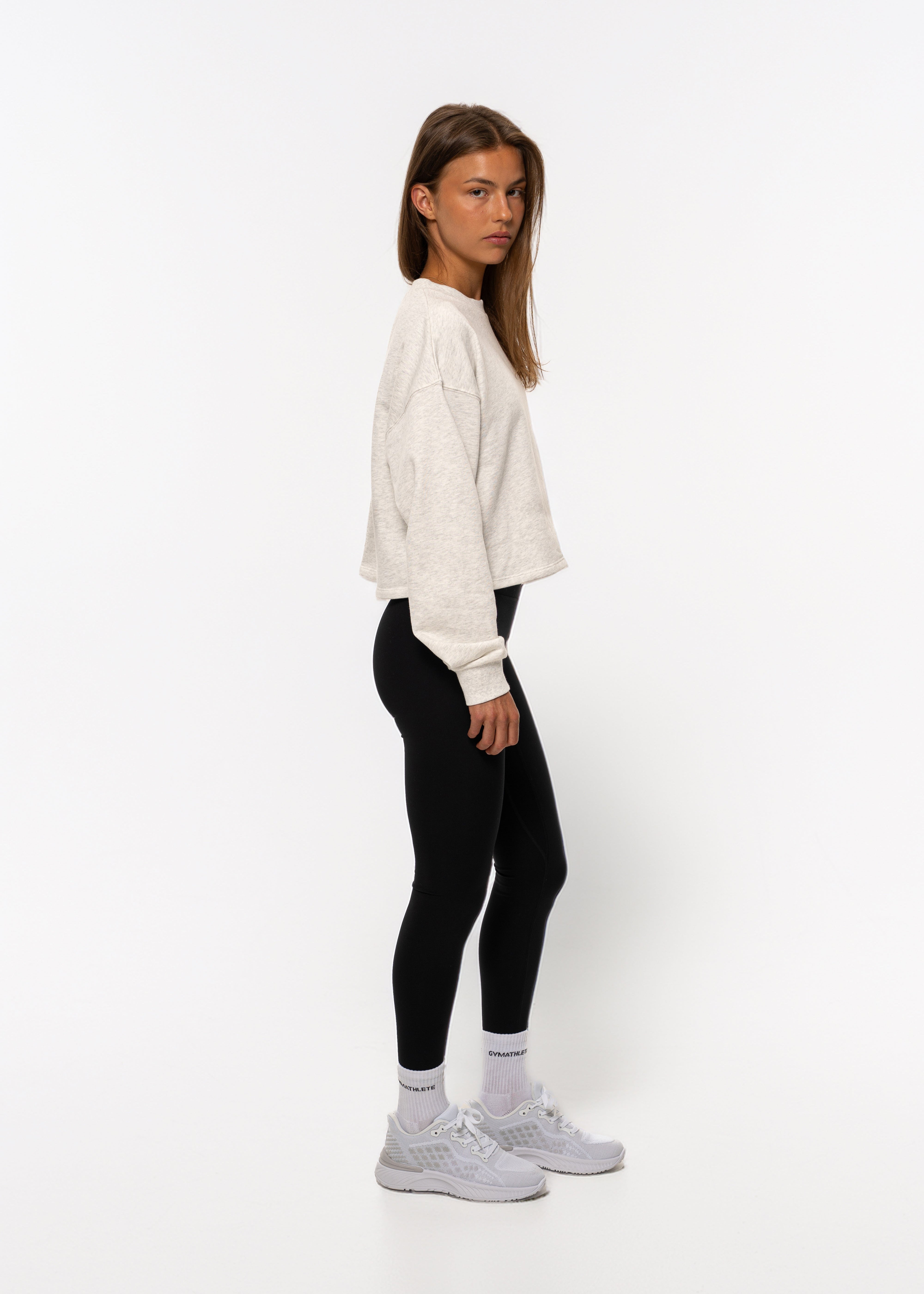 Person wearing a white sweater, black leggings, and white sneakers on a white background. Women - Gymathlete - Flow Crop Sweatshirt - White melange