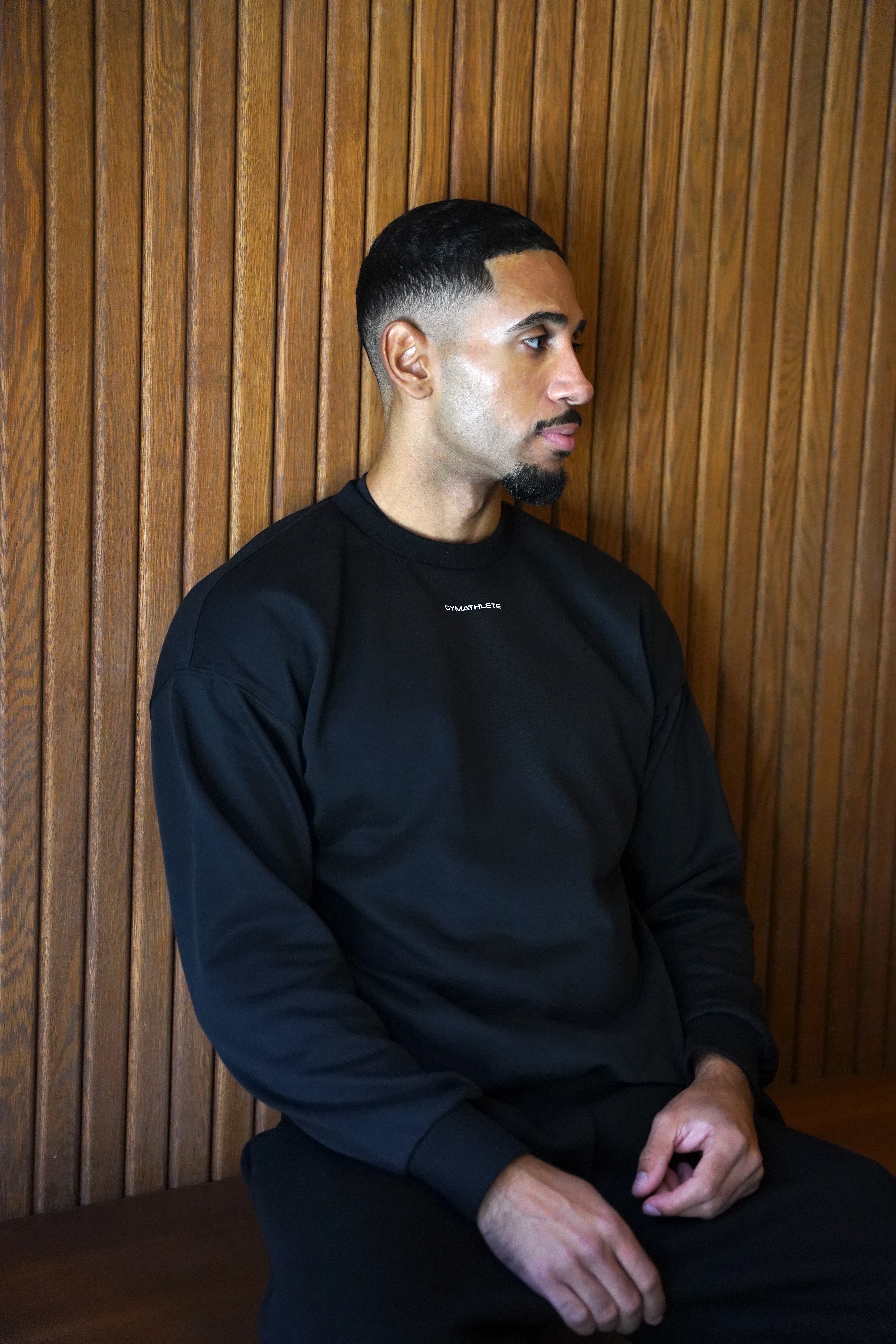 Man wearing a black sweater sitting against a wooden paneled wall. GYMATHLETE. ELEVATE SWEATSHIRT. MEN. GYM. ACTIVEWEAR.