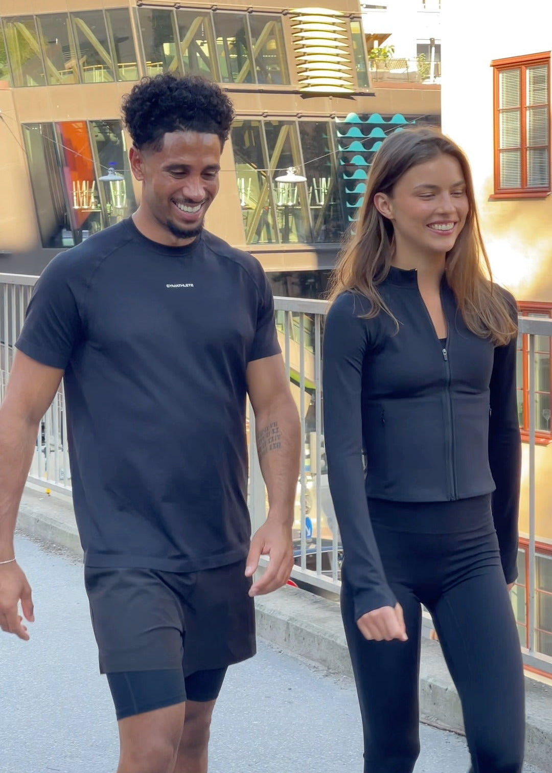 Two people walking outdoors in matching dark athletic wear with a building in the background. - GYMATHLETE