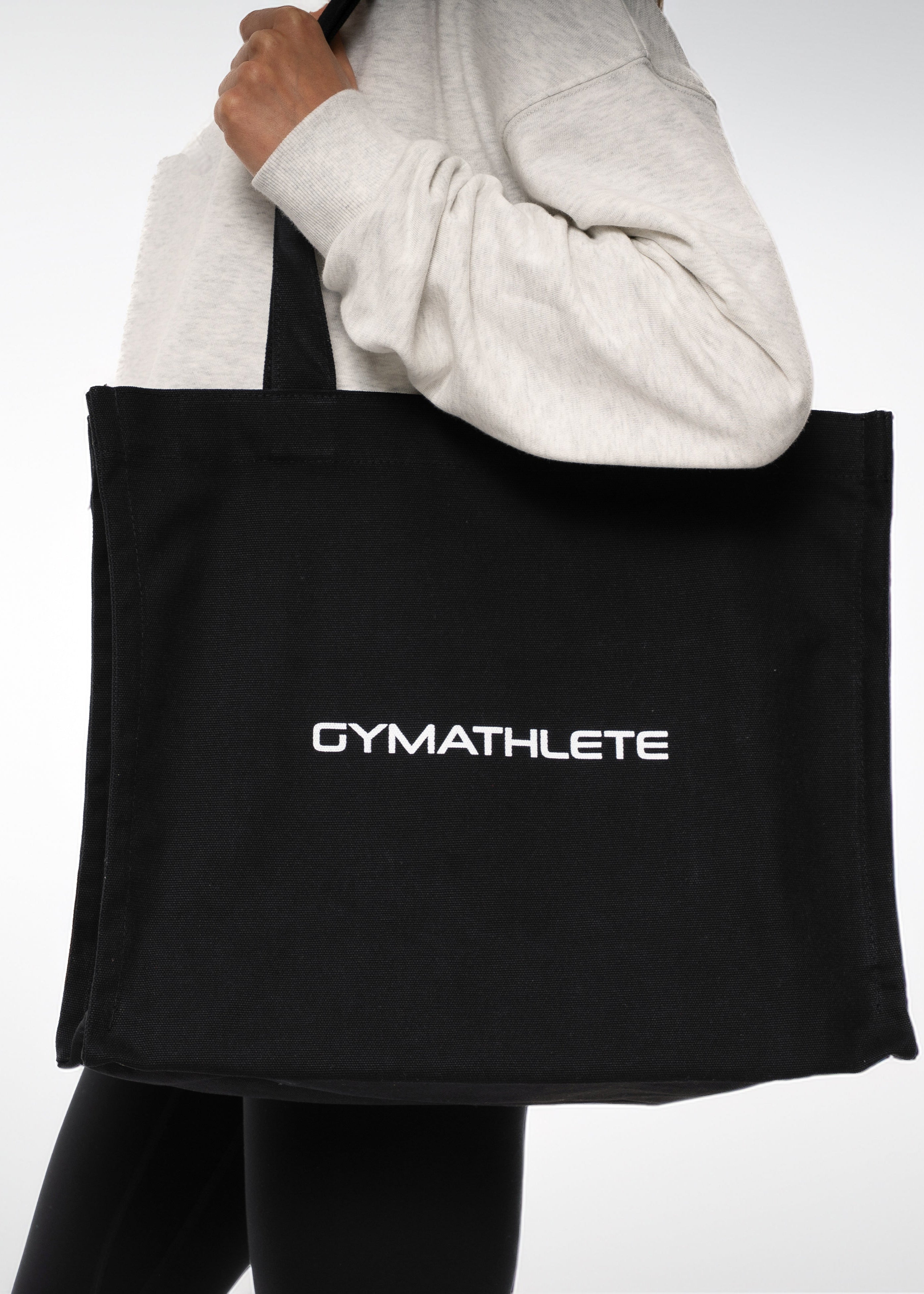 Black tote bag with 'GYMATHLETE' text held by a person wearing a light gray sweatshirt. Gymathlete Balance Tote bag - black