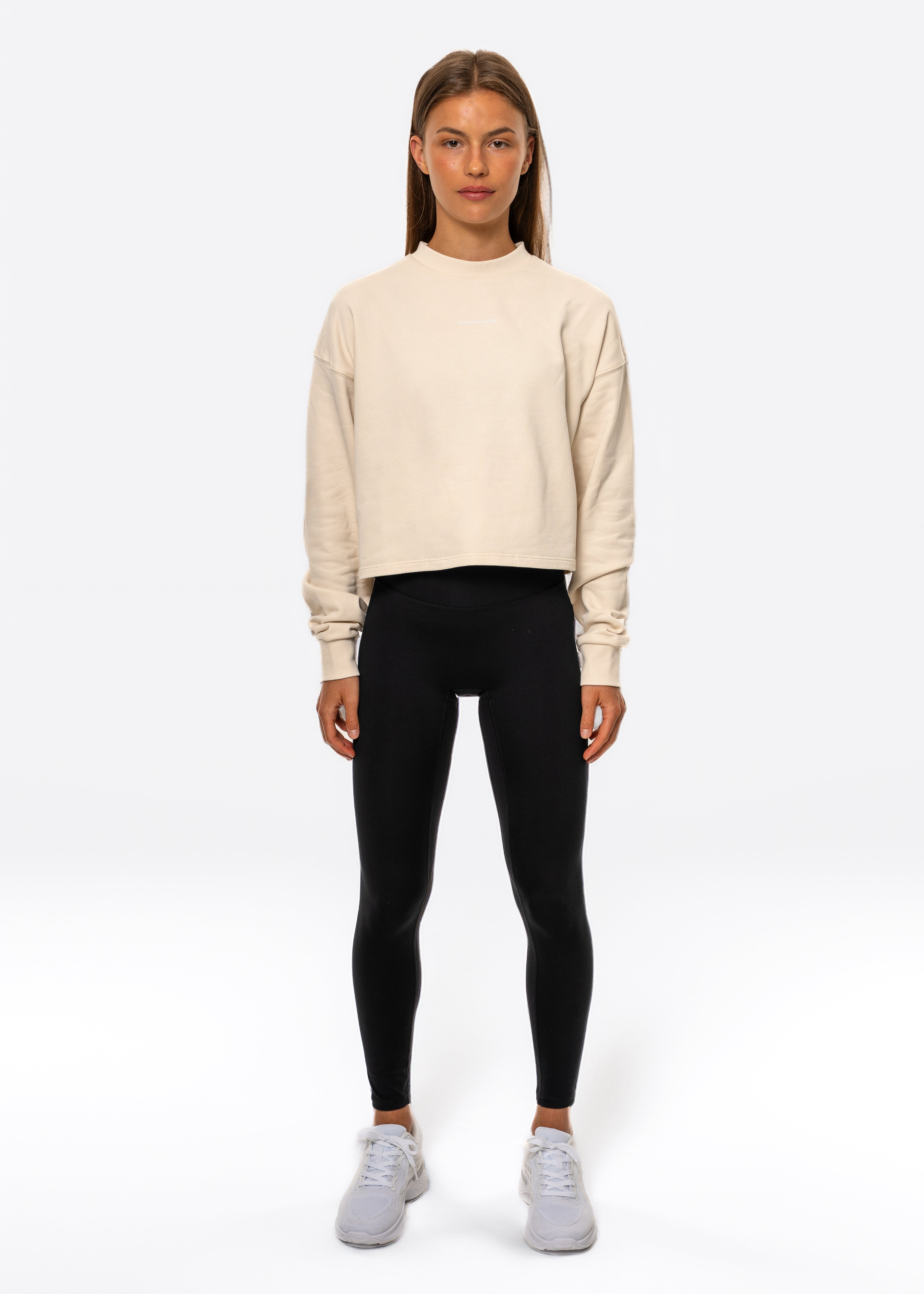 Woman wearing a beige sweater and black leggings on a white background. Gymathlete Flow Crop Sweatshirt. Women.