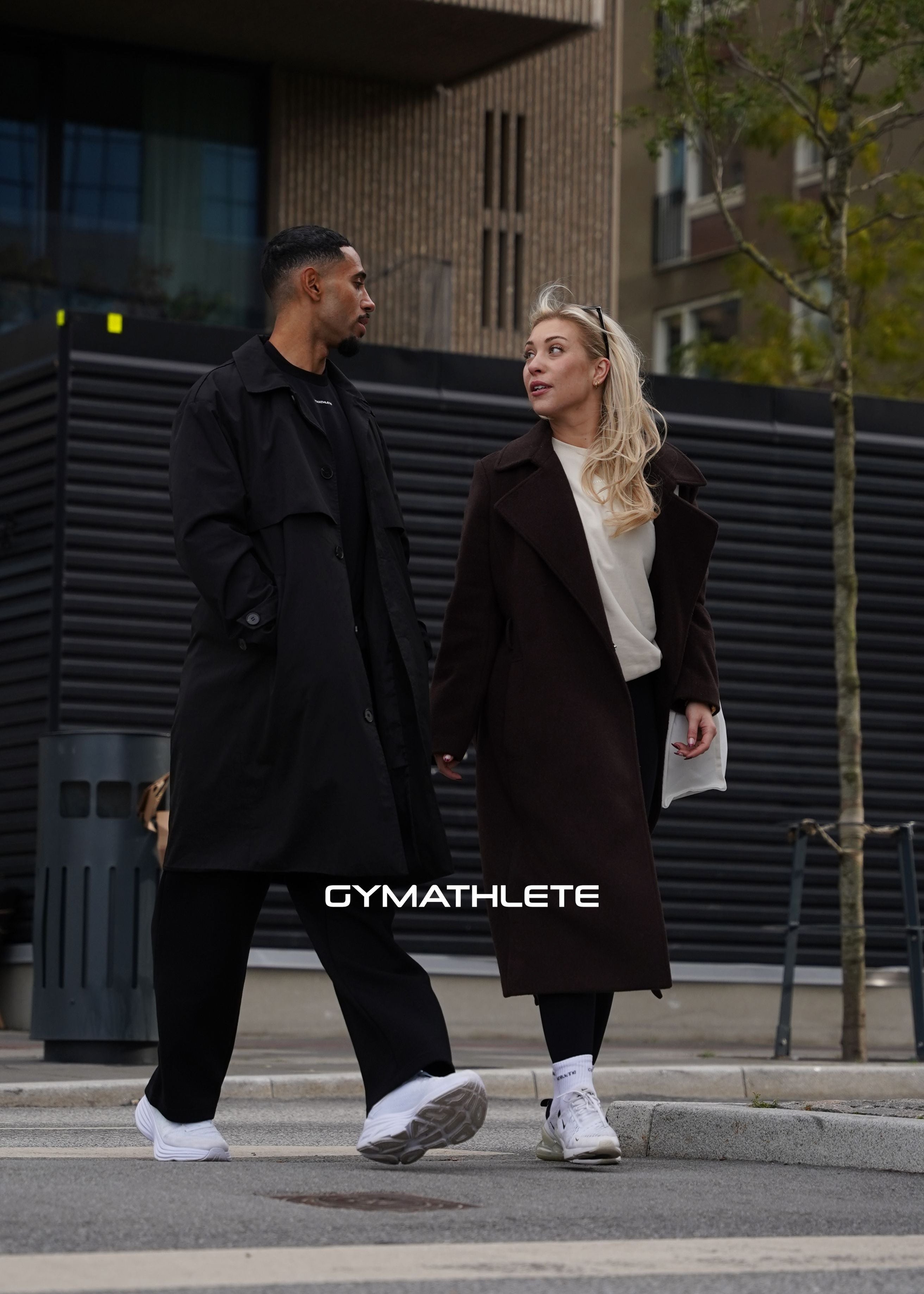 Two people walking on a street, wearing dark coats with 'GYMATHLETE' branding. Women - men - Gymathlete.