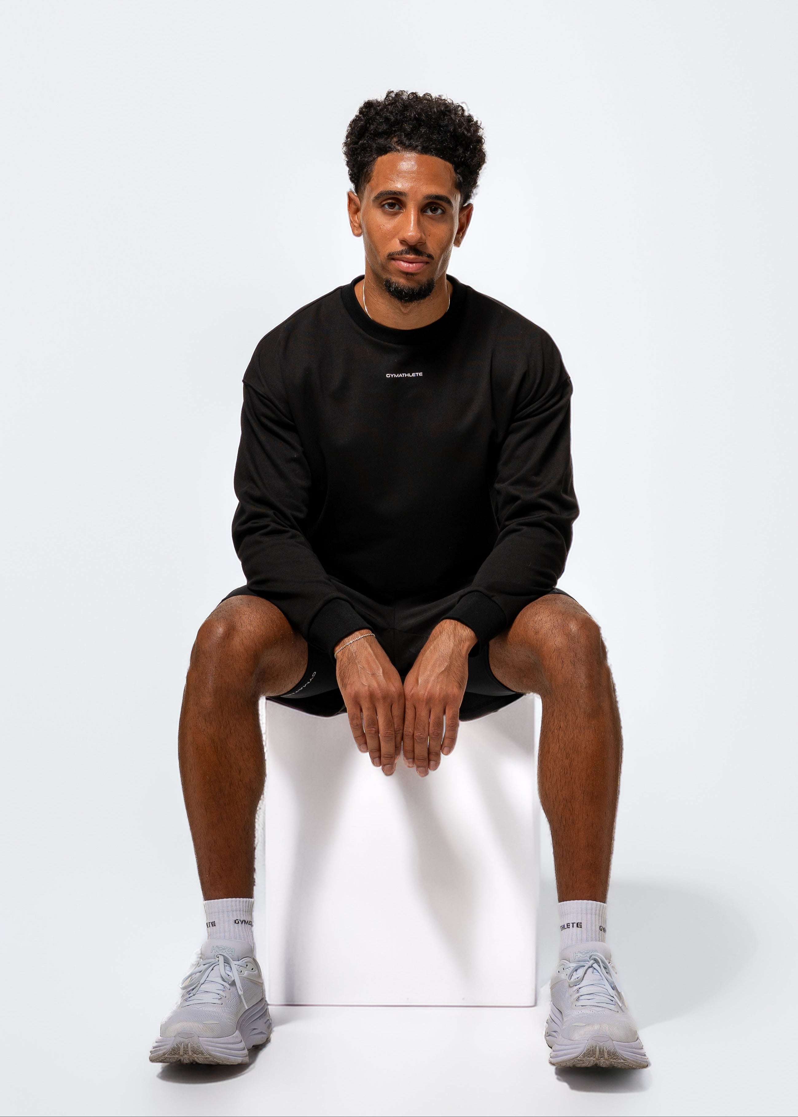 Man sitting on a white stool wearing a black outfit with a visible brand logo on a white background. Gymathlete Endurance socks - men