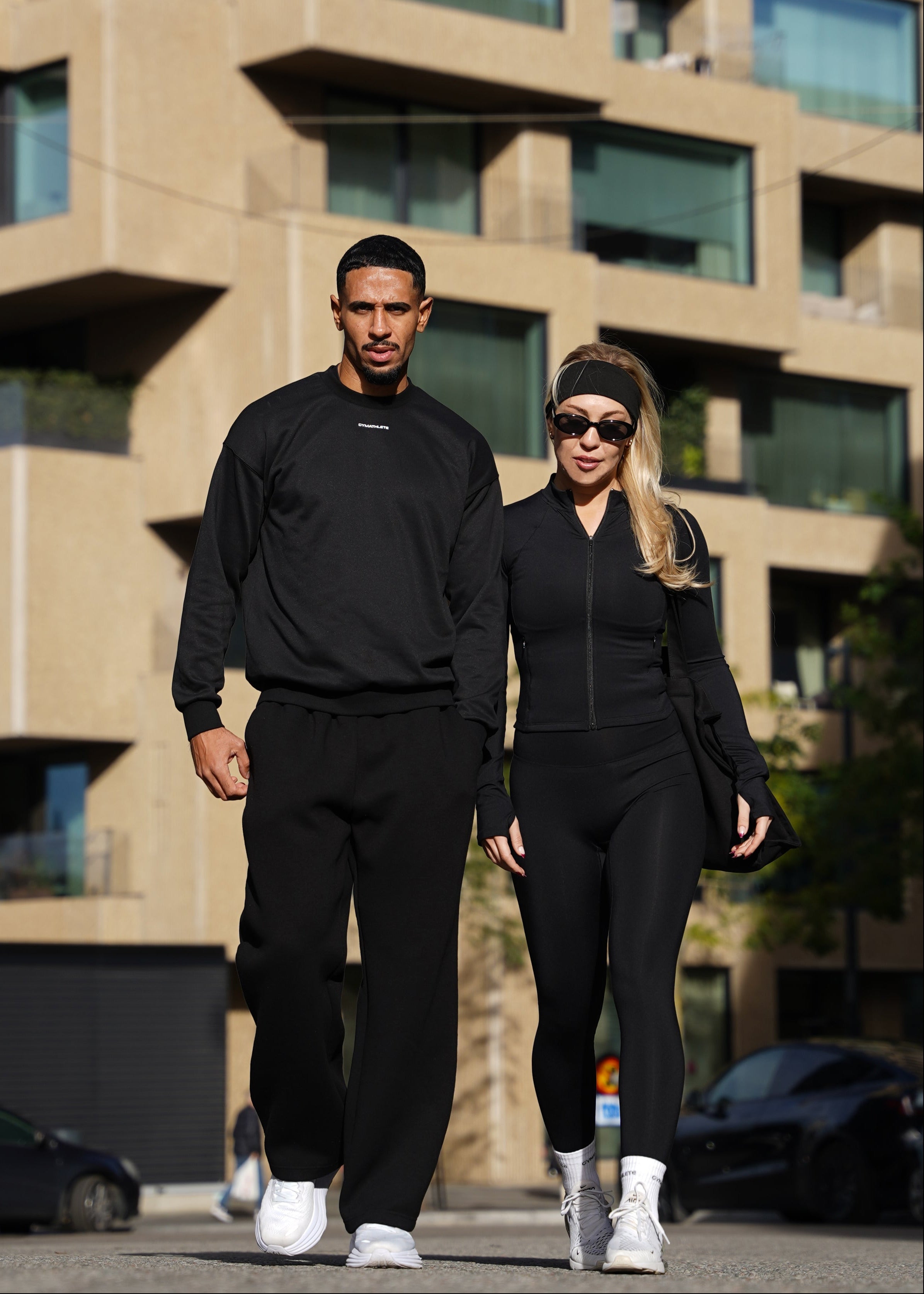 Two people in black outfits walking on a street with a modern building in the background. GYMATHLETE - WOMEN - MEN - GYM - WORKOUT - ACTIVEWEAR