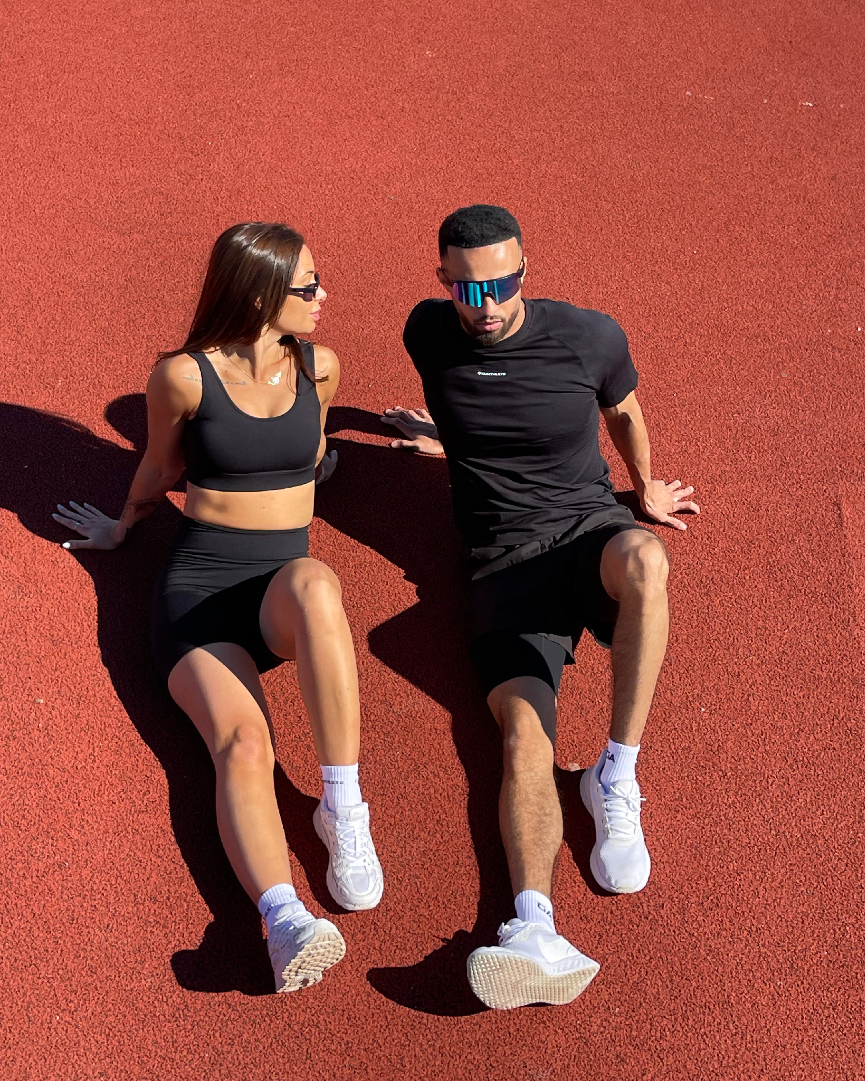 Two athletes in black Gymathlete activewear — a tank top and a technical tee — sitting against a clear blue summer sky, captured for the Summer Workouts and Beyond lookbook.