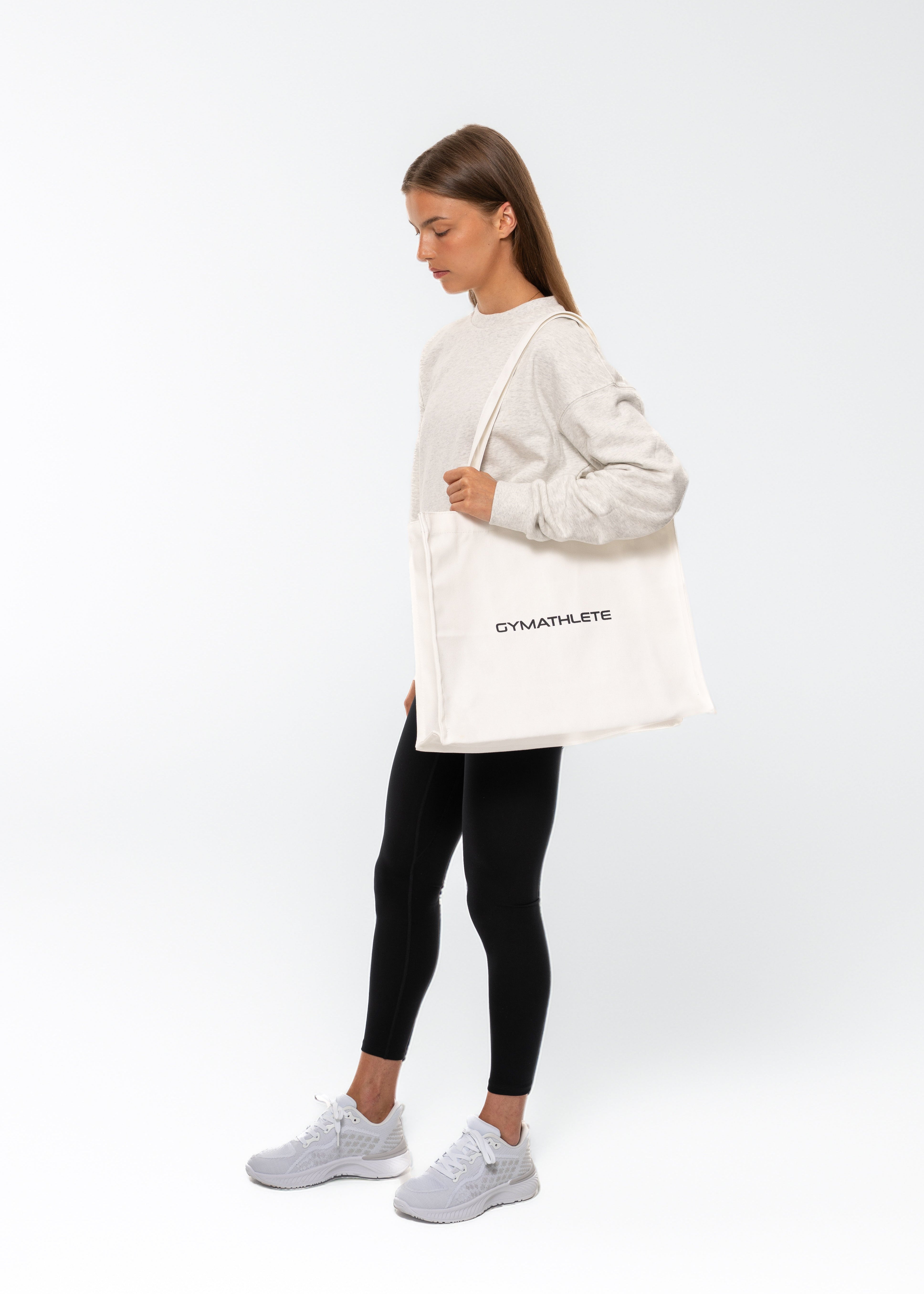 Person holding a white tote bag with text on a white background. 

White tote bag with 'GYMATHLETE' branding on a light gray background. Balance Tote.