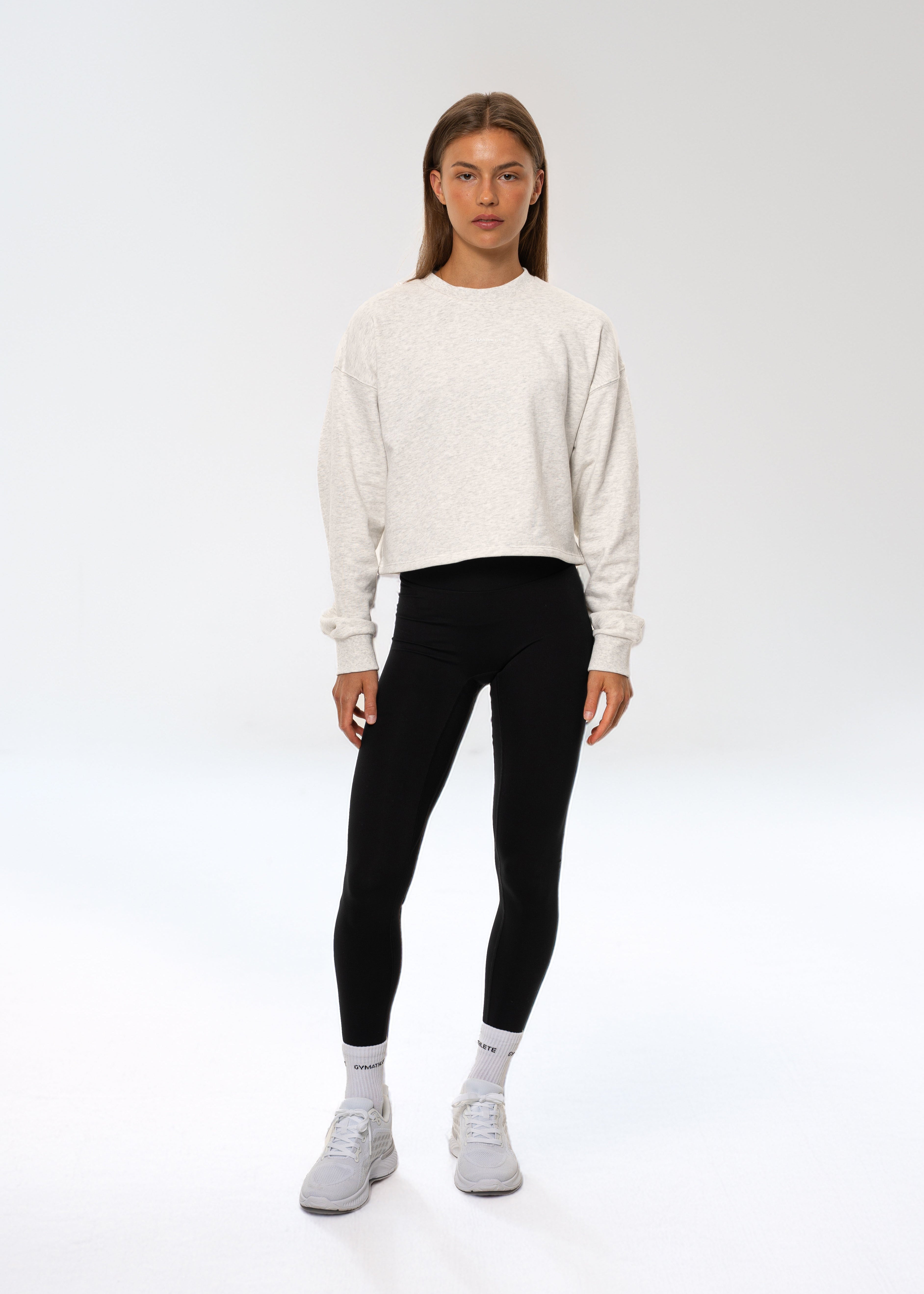 Person wearing a white sweater, black leggings, and gray shoes on a white background. Flow crop sweatshirt, gymathlete