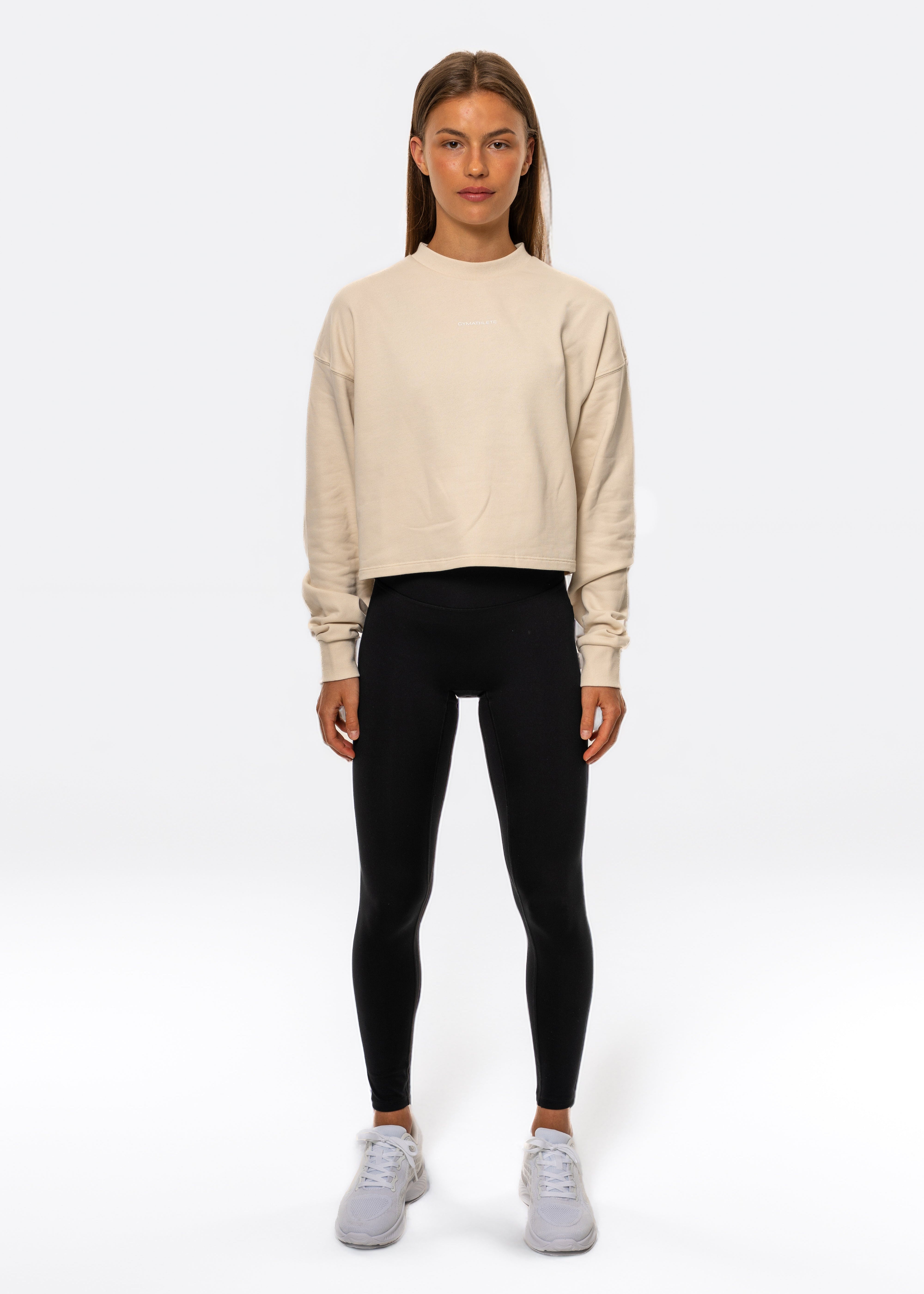 Woman wearing a beige sweater and black leggings on a white background. Gymathlete flow crop sweatshirt