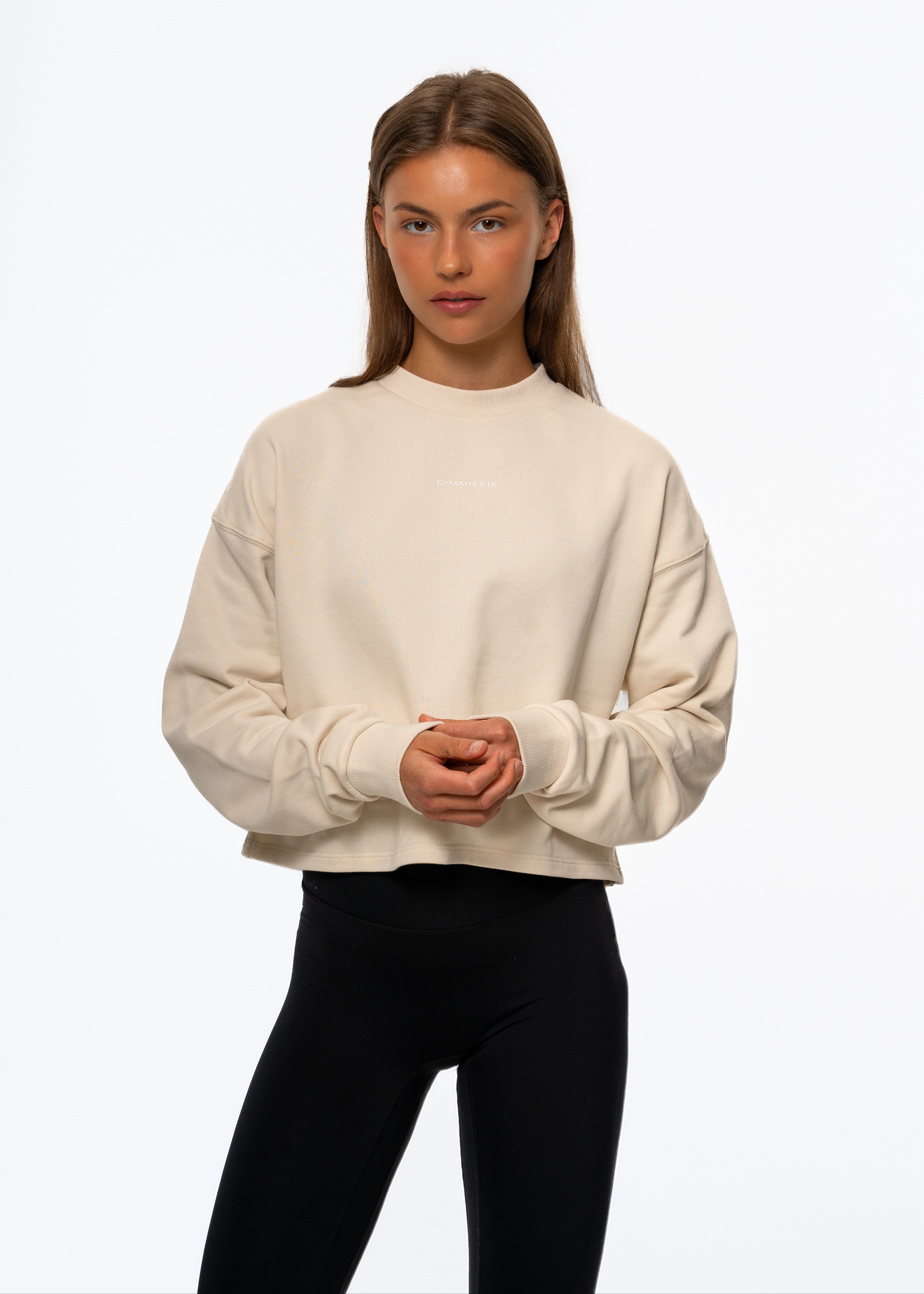 Woman wearing a beige sweater and black pants on a white background. Gymathlete Flow crop sweatshirt