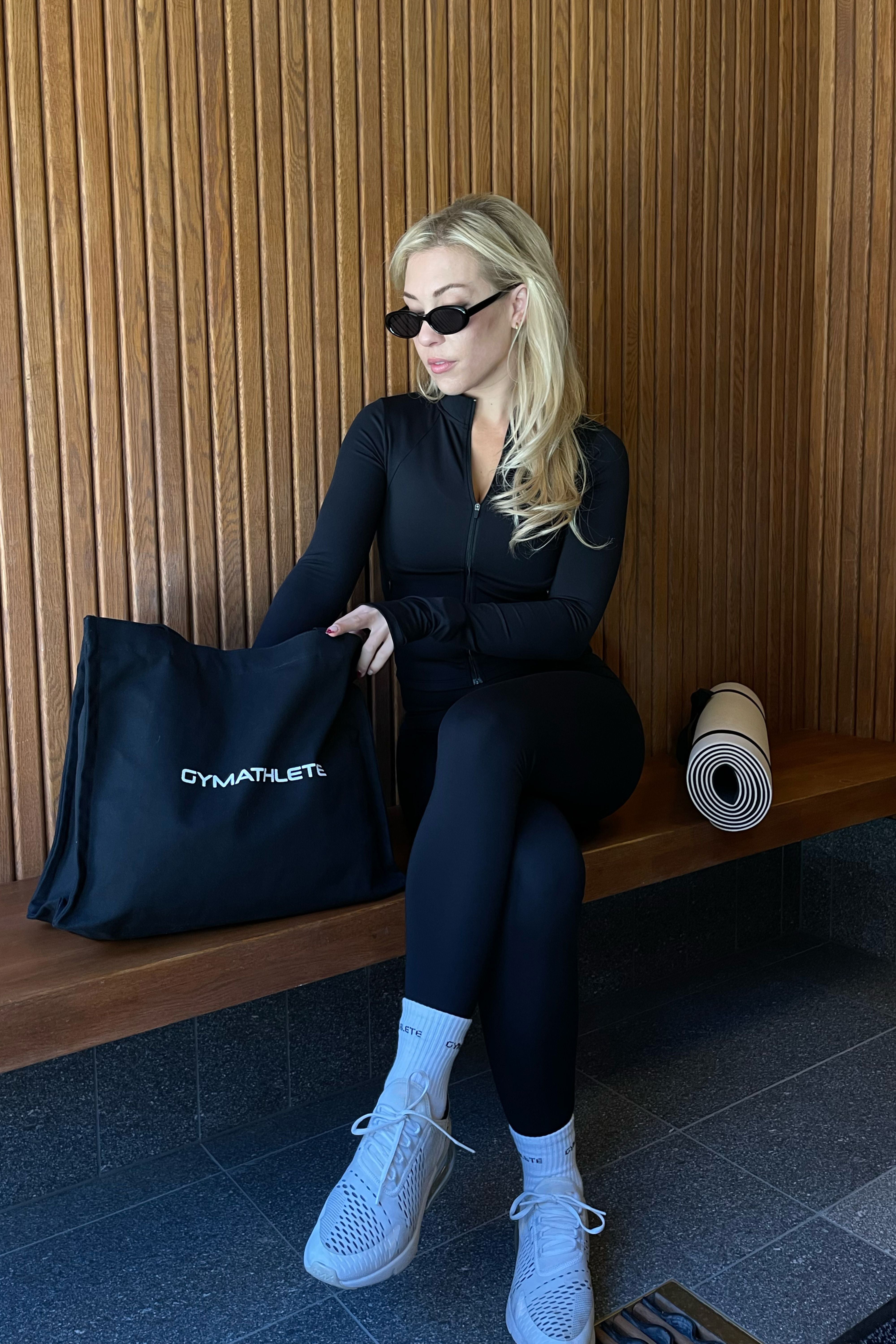 Woman in black athletic wear holding a black tote bag with 'GYMATHLETE' branding, sitting on a wooden bench. Pulse leggings, pulse active jacket, balance tote, endurance socks. Gymathlete.