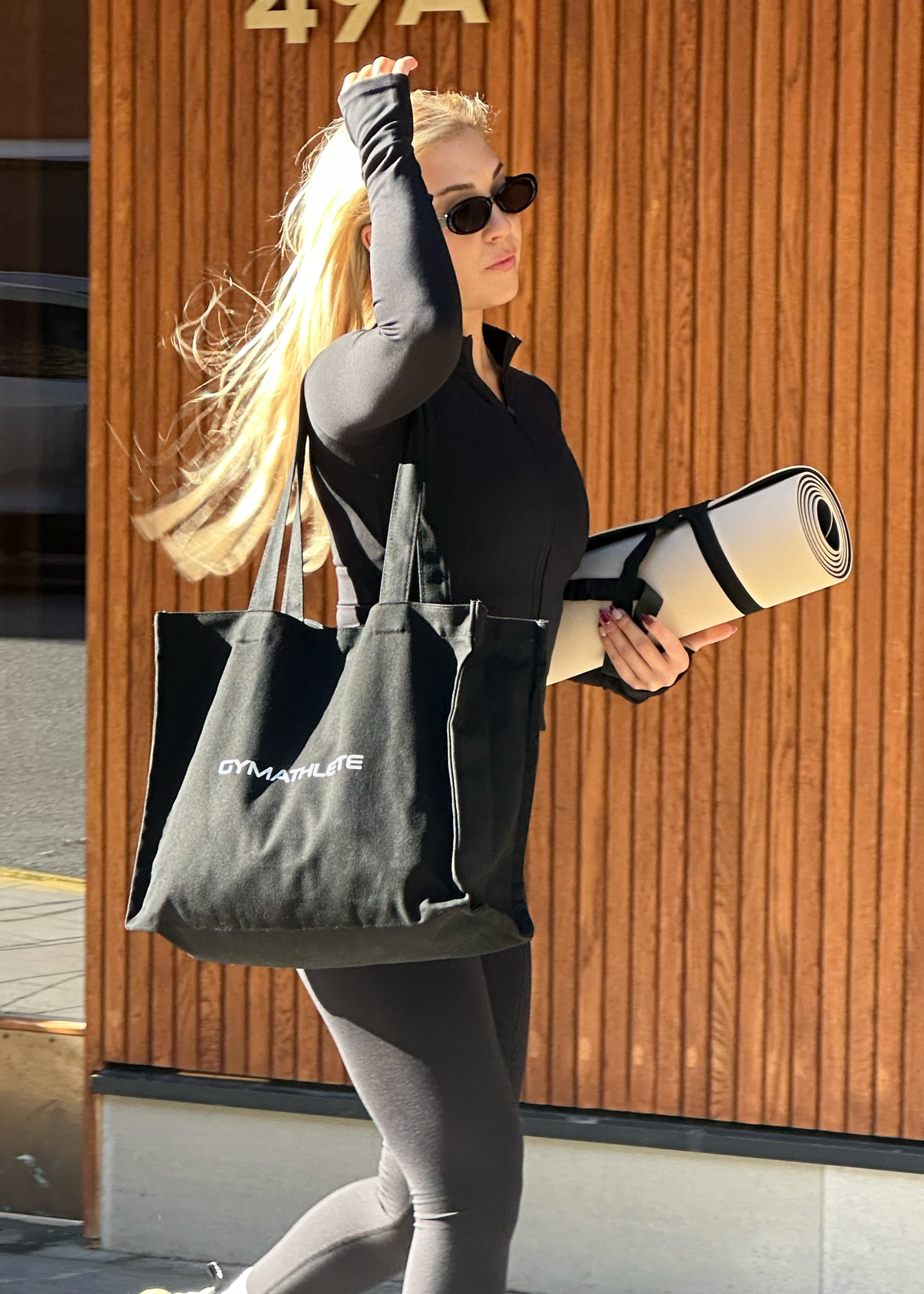 Woman holding a gym mat and a tote bag with 'Gymathlete' branding in front of a wooden door. Gymathlete Balance tote - tote bag.
