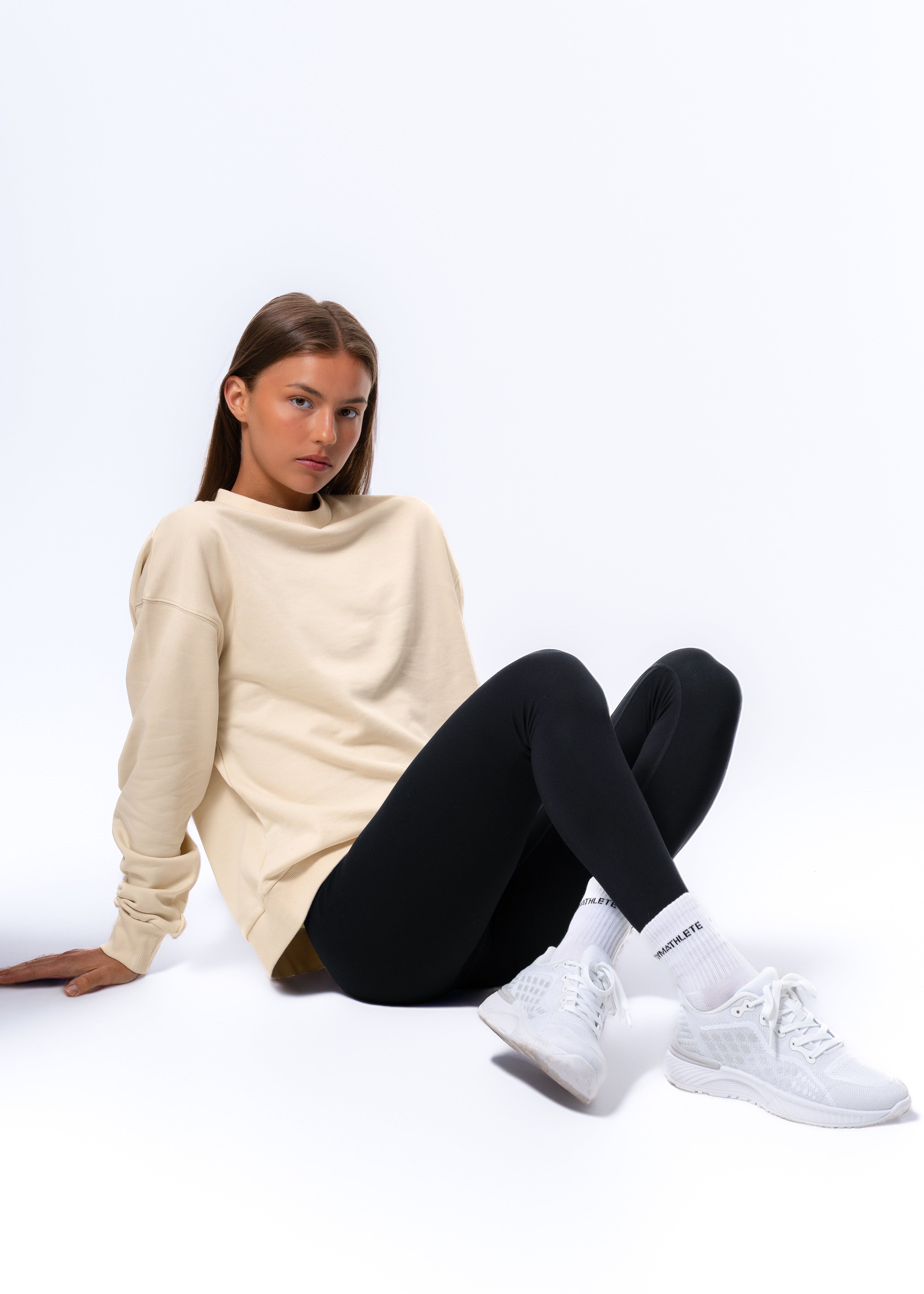 Woman sitting on a white floor wearing a beige sweater, black leggings, and white sneakers. Gymathlete Endurance socks - women
