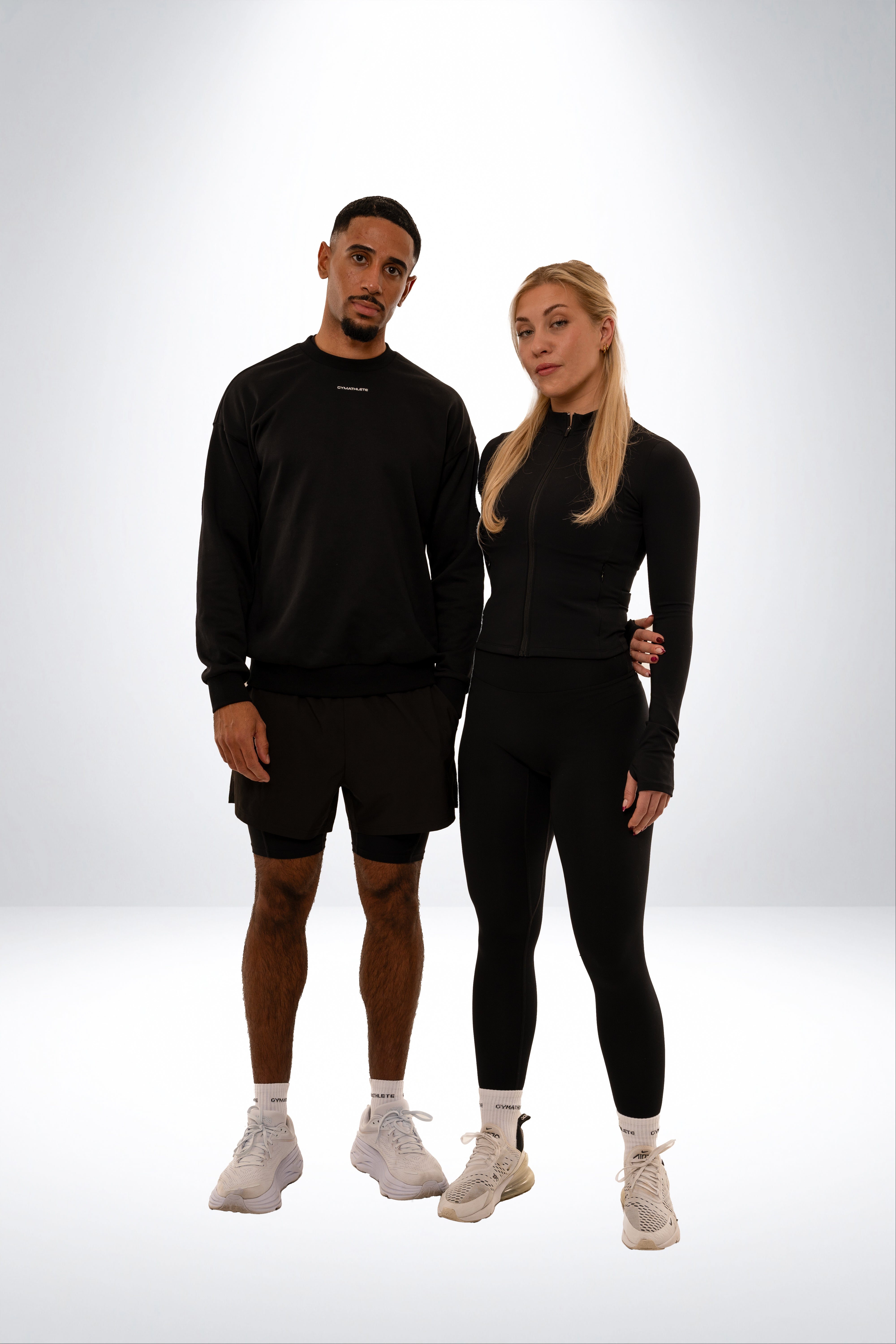 Two people wearing black athletic outfits on a white background. Gymathlete - Elevate Sweatshirt - 2IN1 SHORTS - PULSE LEGGINGS - ENDURANCE SOCKS - ZIP JACKET - Women - men